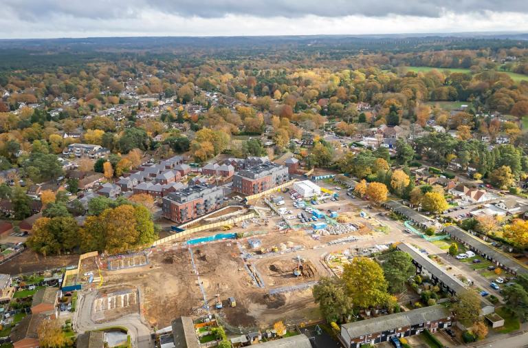 Aerial view of a suburban town with autumn trees and winding streets; foreground shows a construction site with excavators and foundations.