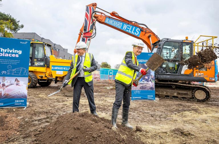 Council and Wates leaders in hard hats shovel dirt at a site with an orange excavator and blue “Thriving places” banners behind.