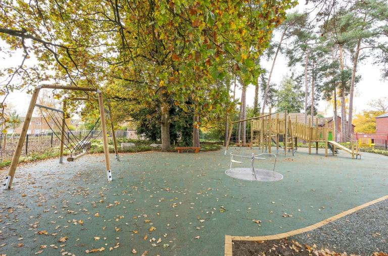 Autumn playground with A-frame swing, climbing frame and slide on green surface with leaves, bench under tree, houses behind fence.