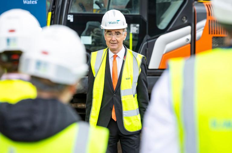 Man in suit with orange tie wears Wates hard hat and safety vest, standing by construction gear with blurred hi-vis workers in front.
