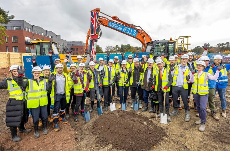 Workers in hi-vis and hard hats celebrate and raise hands on muddy site with an orange excavator bearing a Union Jack behind them.