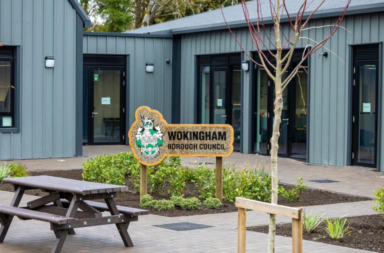 The new grey metal clad office building in the background with picnic benches, newly planted shrubs, trees and a bug hotel sign with Wokingham Borough Council's logo on it