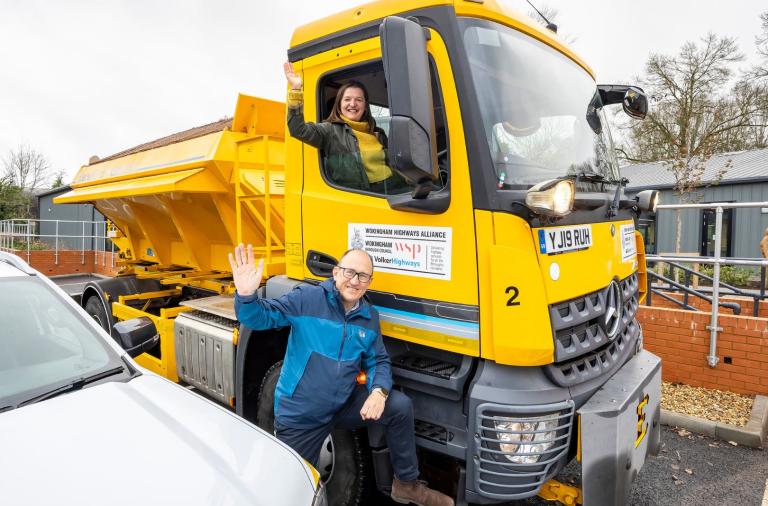 Two people smile and wave at the camera, a lady is in the cab of the gritter, whilst a man has his foot on the step of the gritter cab.