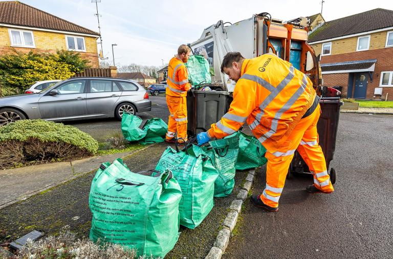 Two waste collectors empty green recycling bags by a truck.