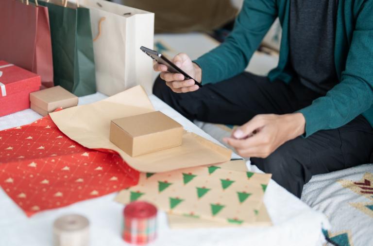 Person using phone near gifts and wrapping paper on a bed.