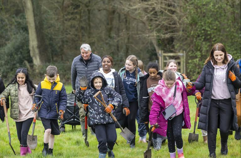 A group of schoolchildren walking across a grassy field carrying spades during a tree planting session at the Covid Memorial Woodland.