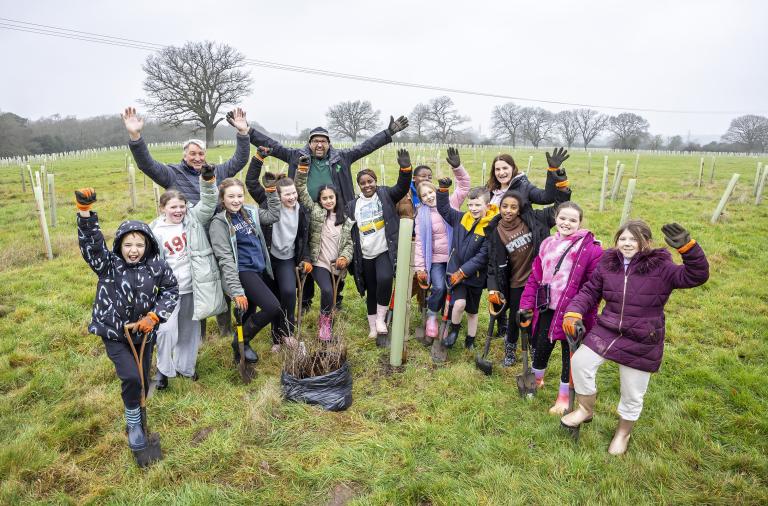 Schoolchildren planting saplings in a field at the Covid Memorial Woodland, holding spades and celebrating their work.