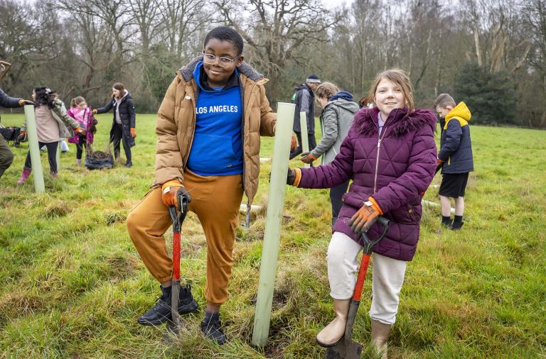 Two children standing beside a newly planted sapling with a tree guard, holding spades during a tree planting session at the Covid Memorial Woodland.