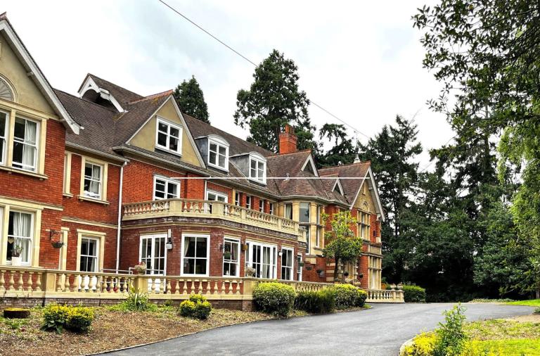 Bluebell House residential home in Wokingham, red brick building with a curved driveway with shrubs leads to a wooded background.