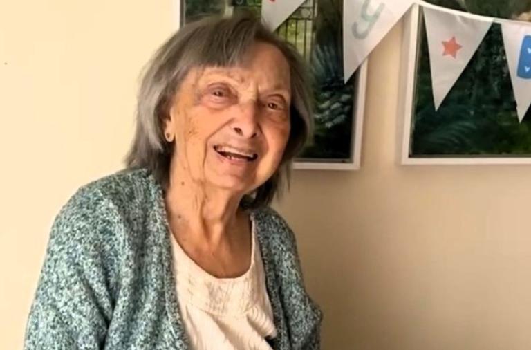 An elderly women with short gray hair, wearing a light patterned cardigan over a white blouse, smiling at the camera