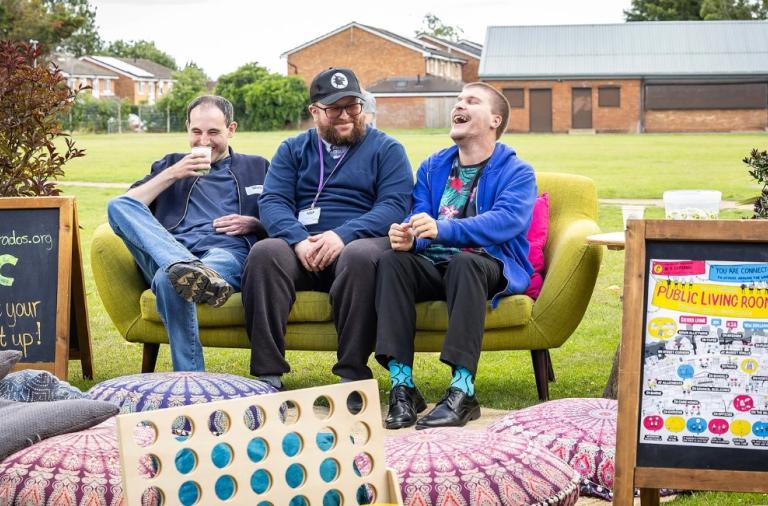 Three men sit on a lime-green sofa on grass, laughing. A chalkboard sign and a Public Living Room poster to each side.