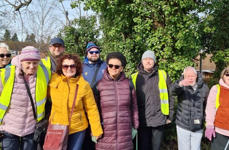 A group of adults wearing bright yellow high-visibility vests pose for a photo outdoors. They stand in front of trees and houses, with several wearing hats, sunglasses, and walking gear.