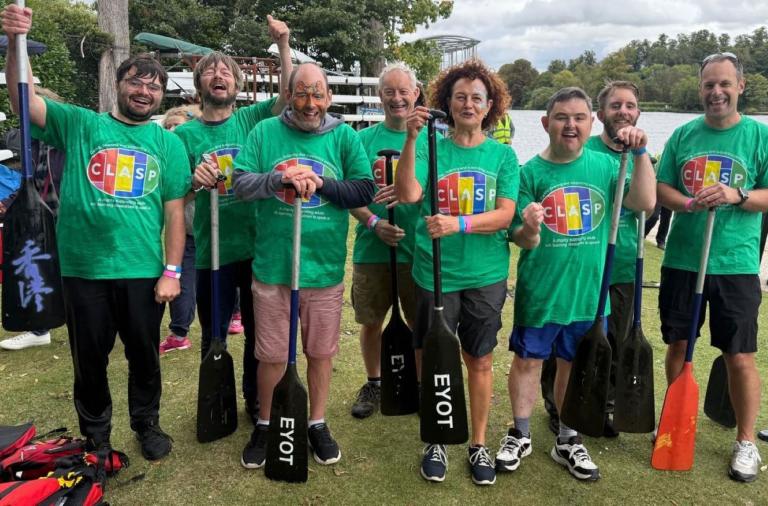 Eight people in matching green CLASP t-shirts stand on grass by a lake, each holding a canoe paddle. They smile and pose for the camera with boats and trees in the background.