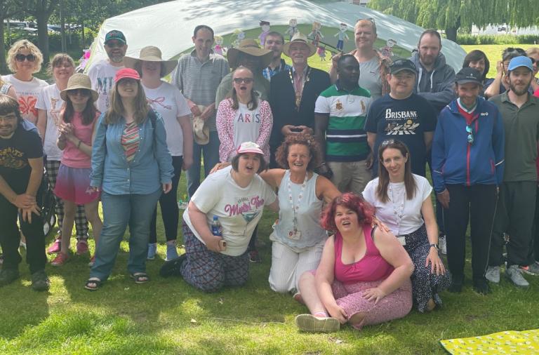 Group of adults posing for a photo outdoors on a sunny, grassy area. They stand and kneel in front of a white canopy tent.