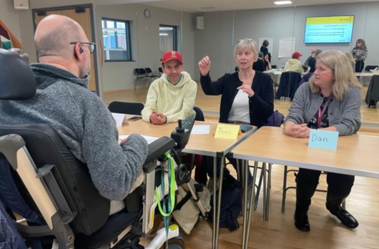 A man in a motorised wheelchair sits at a table with three others in a bright room. A woman in a black jacket gestures while two others listen.