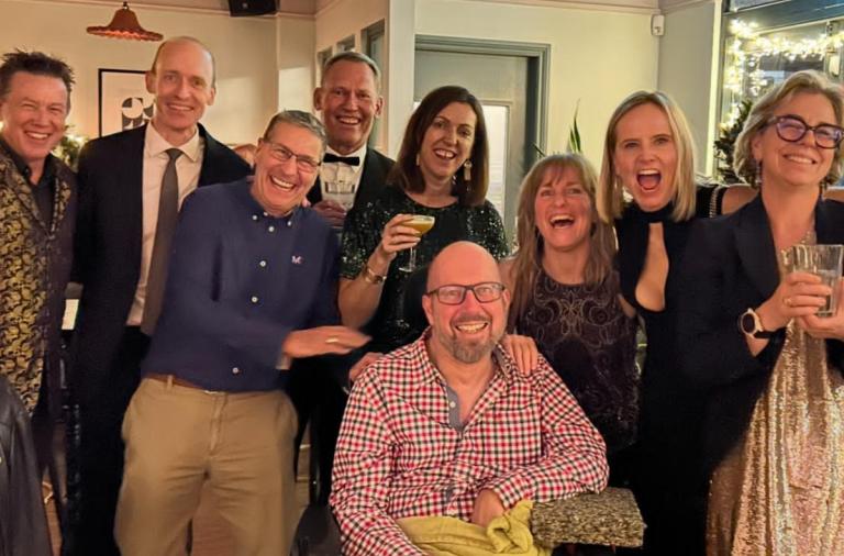 A group of adults posing for a an indoor photo, smiling and holding drinks; a man in a wheelchair sits at the front centre among friends.