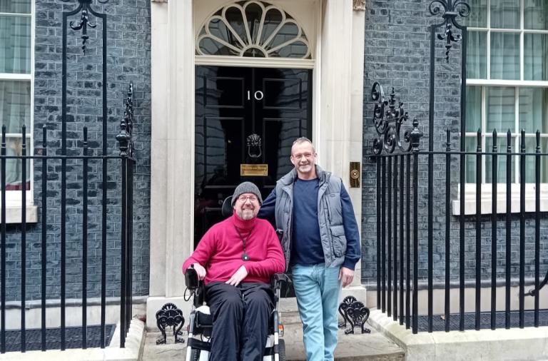 Two men stand outside number 10 Downing Street. One man sits in a wheelchair, while the other stands beside him, with wrought-iron railings along the doorway.