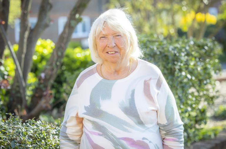 A woman with short white hair stands in a sunny garden, wearing a pastel abstract-patterned sweater. Green shrubs and blurred yellow flowers are in the background.