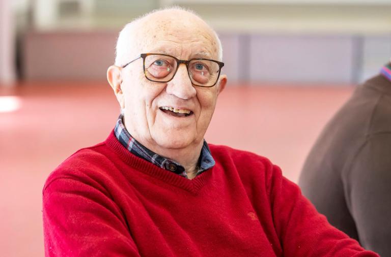 An elderly man with white hair and glasses smiles at the camera. He wears a red sweater over a plaid shirt, and the background is a softly blurred indoor space.