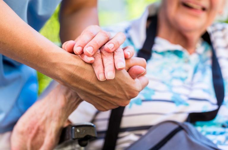 Two hands are clasped in a supportive gesture. In the blurred background, an elderly person wearing a blue floral shirt sits in a wheelchair and smiles.