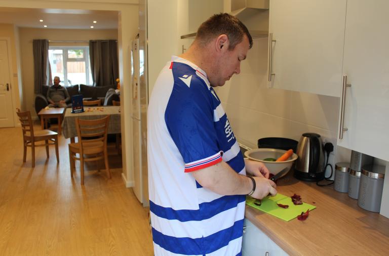 A man wearing a blue and white football shirt stands at a kitchen counter, chopping on a green cutting board. In an adjacent room, another man sits on a sofa near a dining table and chairs.