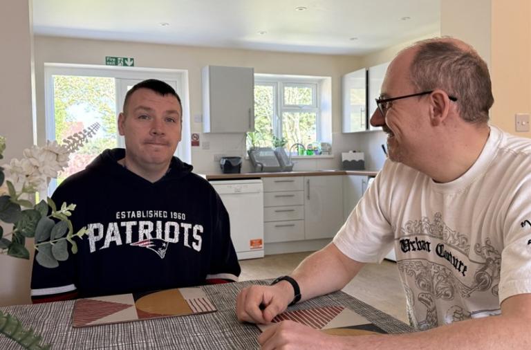 Two men sit across from each other at a dining table in a bright, modern kitchen. The man on the left wears a black Patriots hoodie; the man on the right wears a white graphic T‑shirt and glasses, and they smile at each other.