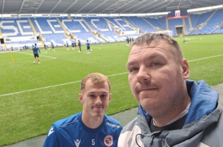 Two men take a selfie on a football pitch inside Reading stadium. The man in front wears a blue jacket, while the man behind wears a blue training top with the number 5, with players training in the background.