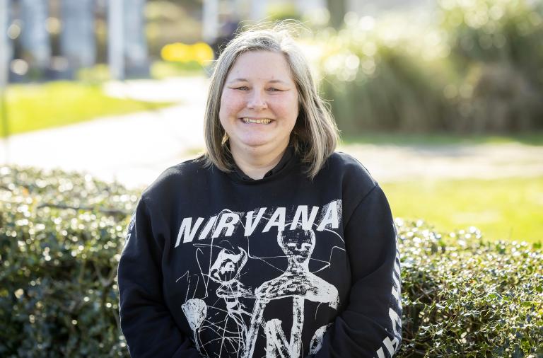 A person with shoulder-length blond hair smiles outdoors. They wear a black Nirvana sweatshirt with a skeletal graphic, standing in front of hedges on a sunny day.