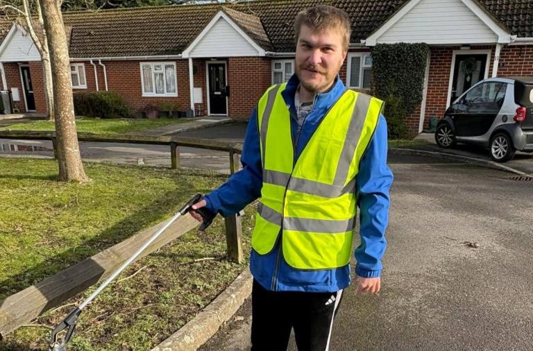 A man in a high-visibility vest over a blue jacket stands on a street, holding a long-handled litter picker. Brick houses, a hedge, and a parked car are in the background.