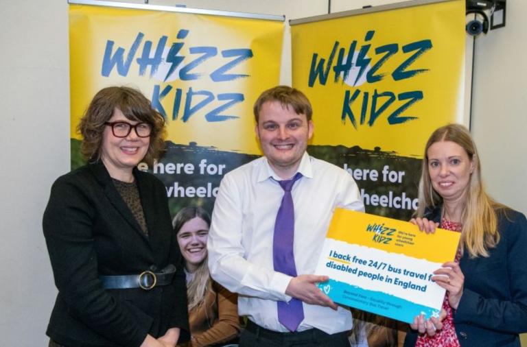 Three people pose in front of yellow Whizz-Kidz banners. The centre person in a white shirt and purple tie is flanked by a woman with glasses on the left and a woman on the right holding a sign supporting free 24/7 bus travel for disabled people in England.