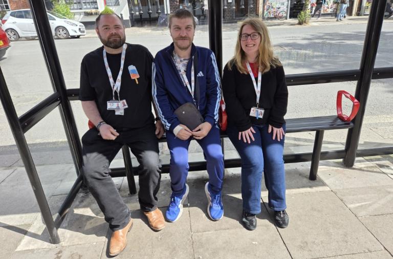 Three people sit on a bench inside a glass bus stop shelter on a town street. Left to right: a bearded man in a black T-shirt and tan boots, a man in a blue tracksuit holding a dark folder, and a smiling woman with glasses in a black jumper and jeans.