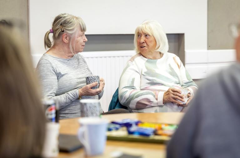 Two older women sit at a table in a bright room, chatting; a tray of snacks and cups is on the table. The left woman has grey hair in a ponytail, while the right woman with white hair wears a pastel jumper and holds a mug.