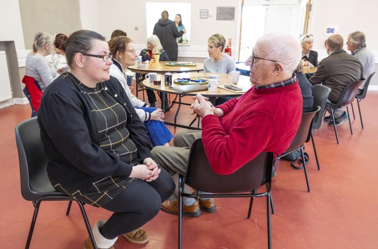 Two people sit facing each other at a round table in a bright community room, a woman in a black cardigan and check dress chatting with an elderly man in a red jumper. In the background, other groups sit at tables.