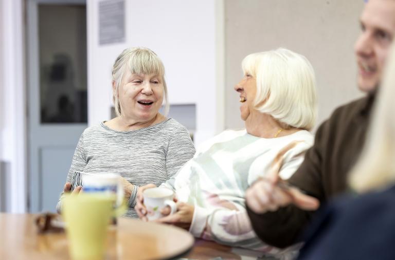 Three older adults sit at a round table, smiling and holding mugs. One has grey hair and a striped top, another has white hair and a pastel jumper; a fourth person is partly seen on the right.