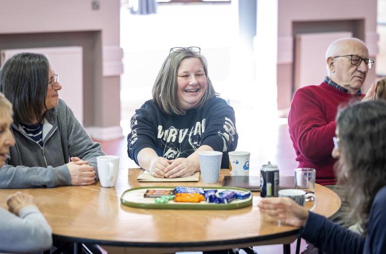 Adults sit around a round wooden table in a bright room, smiling. Mugs, a can, a notebook, and a tray of assorted snacks sit on the table.