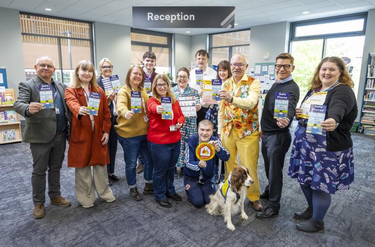 A group of adults pose in a library reception area, all holding blue 'Safe Places' leaflets. A dog in a vest sits in front; a 'Reception' sign and windows are in the background.