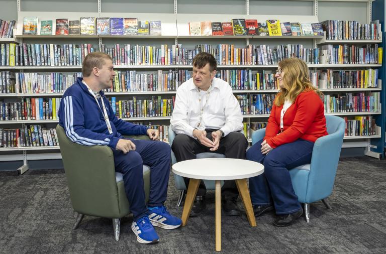 Three people sit in armchairs around a small white table in a library. Shelves of books line the background; left in a blue tracksuit, middle in a white shirt, right in a red cardigan.