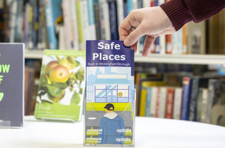A hand in a maroon sleeve holds a vertical leaflet titled Safe Places, showing a building and a person in a blue jacket; it rests on a white table with bookshelves in the background.