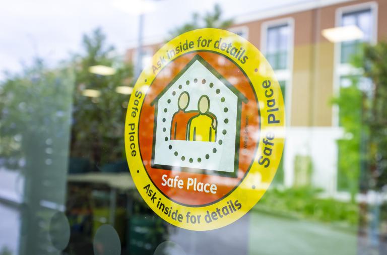 A circular yellow sticker on a window reads “Safe Place” with the text “Ask inside for details” around the edge. Inside is a white house icon with two abstract figures in orange and yellow, framed by a dotted border, with reflections of a building and greenery in the glass.