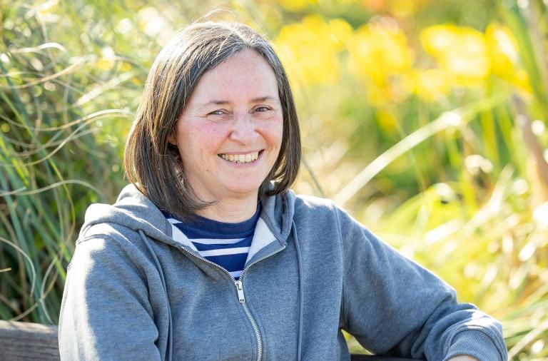 Smiling woman with short dark bob, wearing a blue striped shirt under a grey zip hoodie. Outdoors, sunlit greenery provides a blurred yellow-green background.