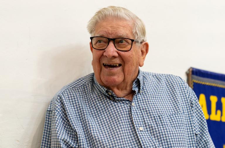 An elderly man with white hair and black-framed glasses smiles. He wears a blue-and-white checkered shirt; a blue banner with yellow letters is visible in the background on the side.