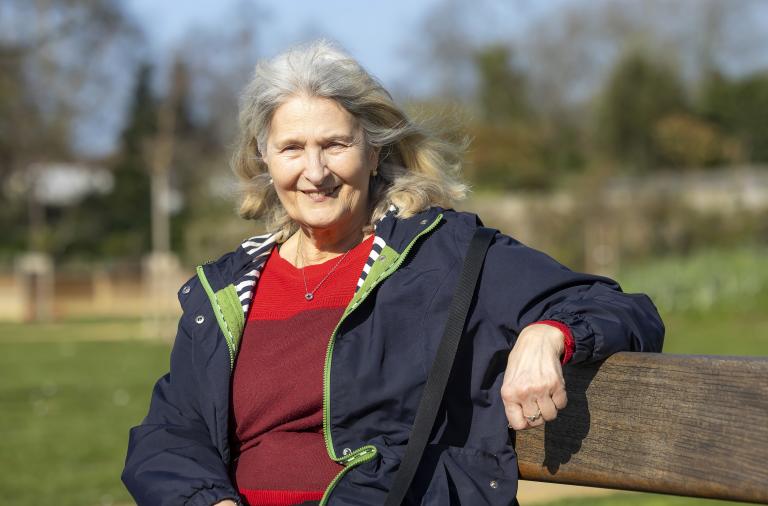 An older woman with grey hair sits on a wooden park bench, smiling at the camera. She wears a navy jacket over a red jumper, with a bright green zipper and a crossbody bag, in a sunny park with blurred trees in the background.