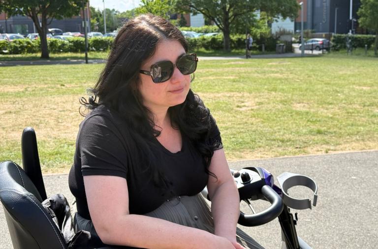 A woman with long dark hair wearing large sunglasses and a black T-shirt sits on a mobility scooter in a sunny park. In the background, there is grass, trees, and buildings with parked cars along a street.