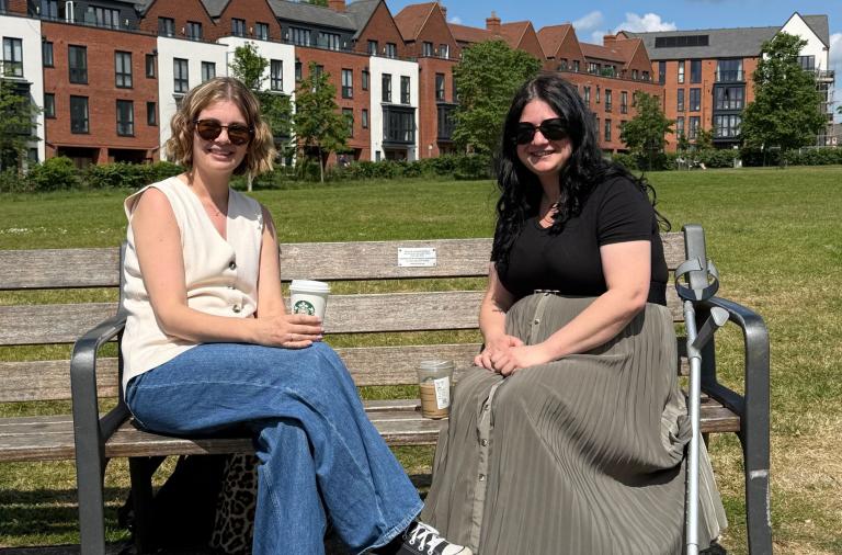 Two women wearing sunglasses sit on a wooden park bench on a sunny day, the left in a cream sleeveless top and jeans holding a coffee cup, the right in a black top and grey skirt beside a mobility aid. Red-brick apartment buildings and a green lawn rise behind them.