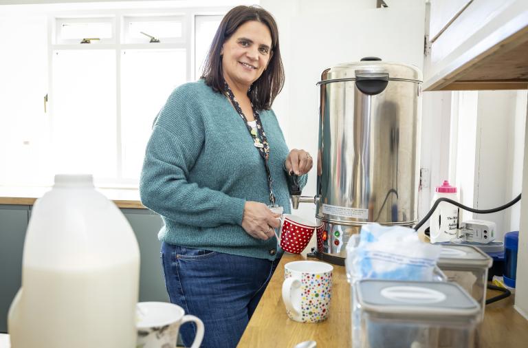 A woman in a teal jumper stands at a kitchen counter beside a large stainless steel coffee urn, smiling and holding a red polka-dot cup. Mugs, a milk jug, and various supplies sit nearby, with windows in the background.