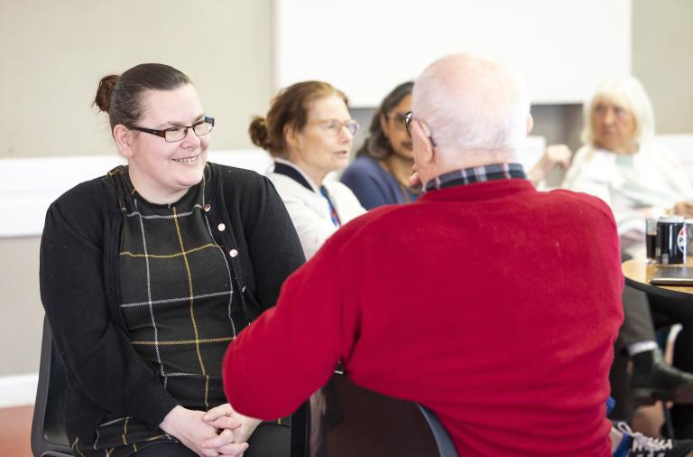 A woman with dark hair, glasses, and a black cardigan over a plaid dress sits smiling as she talks to an older man in a red sweater. In the background, several people sit around a table with mugs.