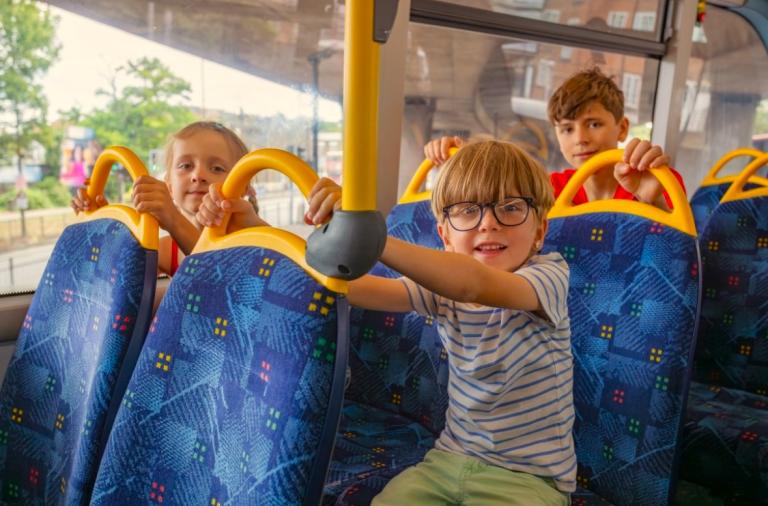 Two young boys and one young girl sitting on the back seats of a bus while smiling at camera