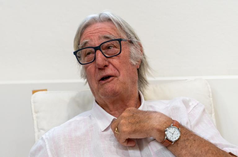An older man with grey hair and dark rectangular glasses sits on a light chair against a pale wall. He wears a light shirt and a brown leather wristwatch on his left wrist, and he appears to be speaking.
