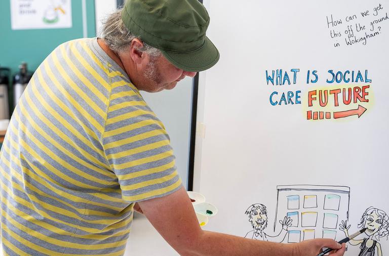 A man in a green cap and yellow-striped shirt writes on a whiteboard. The artwork shows the title “What is social care future” in blue and red with an orange arrow, plus a handwritten question about starting it in Wokingham and cartoon figures at the bottom.