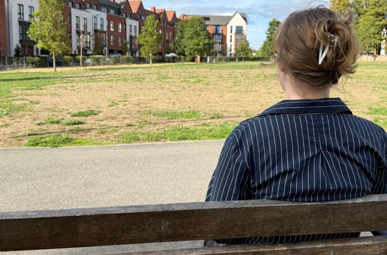 Back view of a person with light brown hair tied with a hair clip, wearing a navy pinstriped shirt, seated on a wooden bench in an urban park.